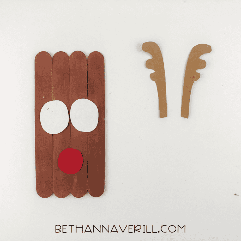 A red paper nose attached below the white paper eyes on the brown craft stick reindeer face.