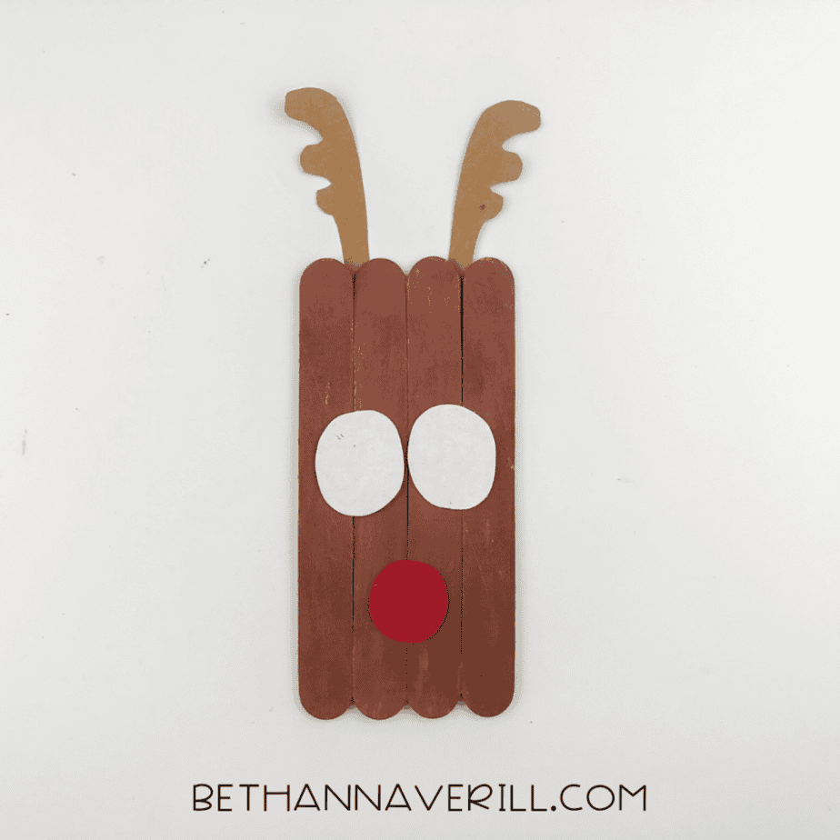 A reindeer craft made from brown-painted craft sticks with attached antlers, white eyes, and a red nose on a white background.