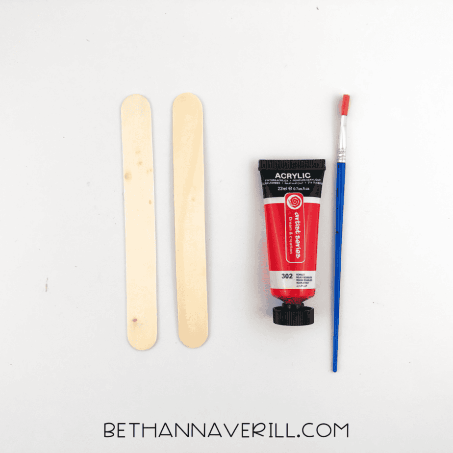 Two plain wooden popsicle sticks placed beside red acrylic paint and a paintbrush on a white background.