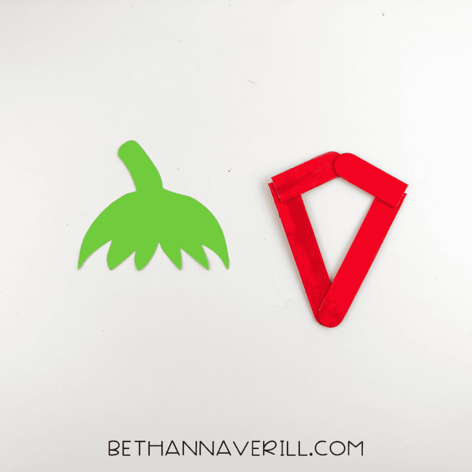 A green paper strawberry top with leafy edges placed next to the popsicle stick frame.