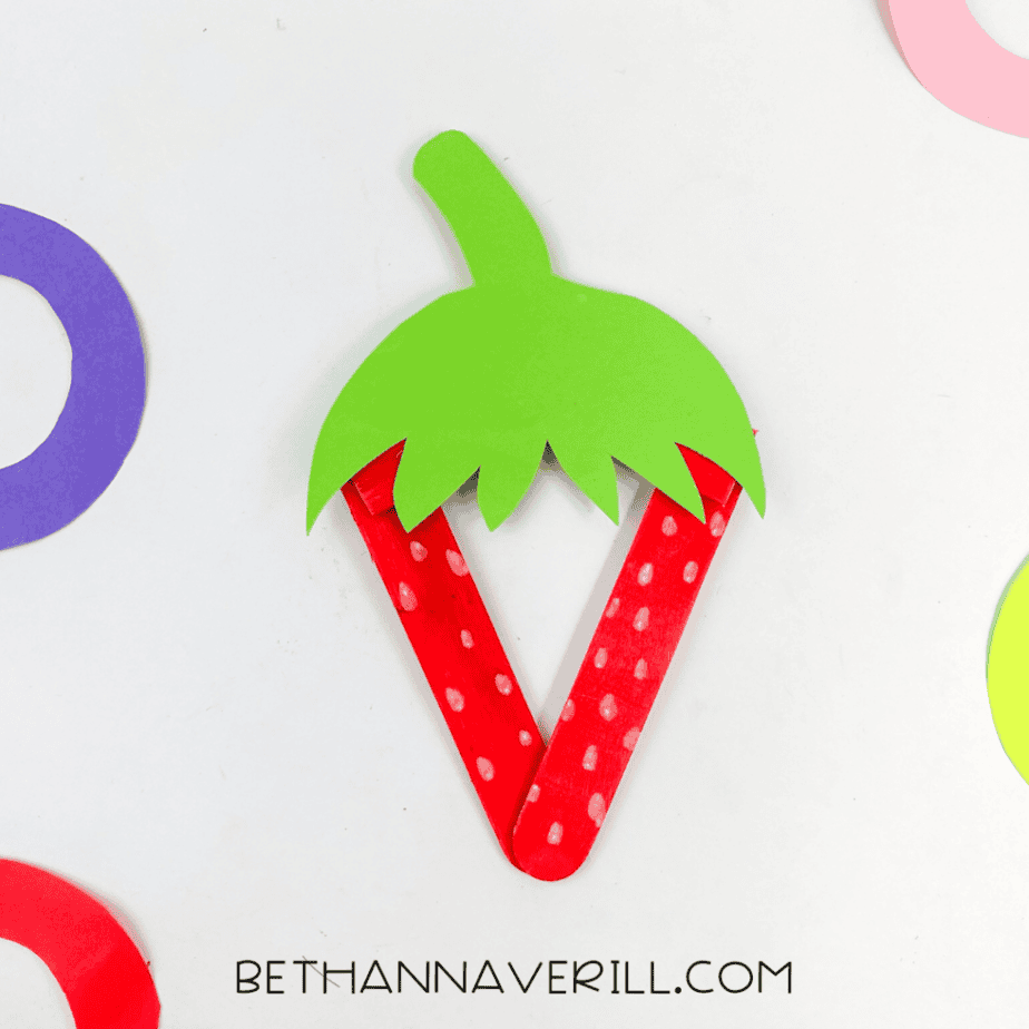 The finished strawberry popsicle stick craft displayed on a white background with colorful paper shapes partially visible around it.