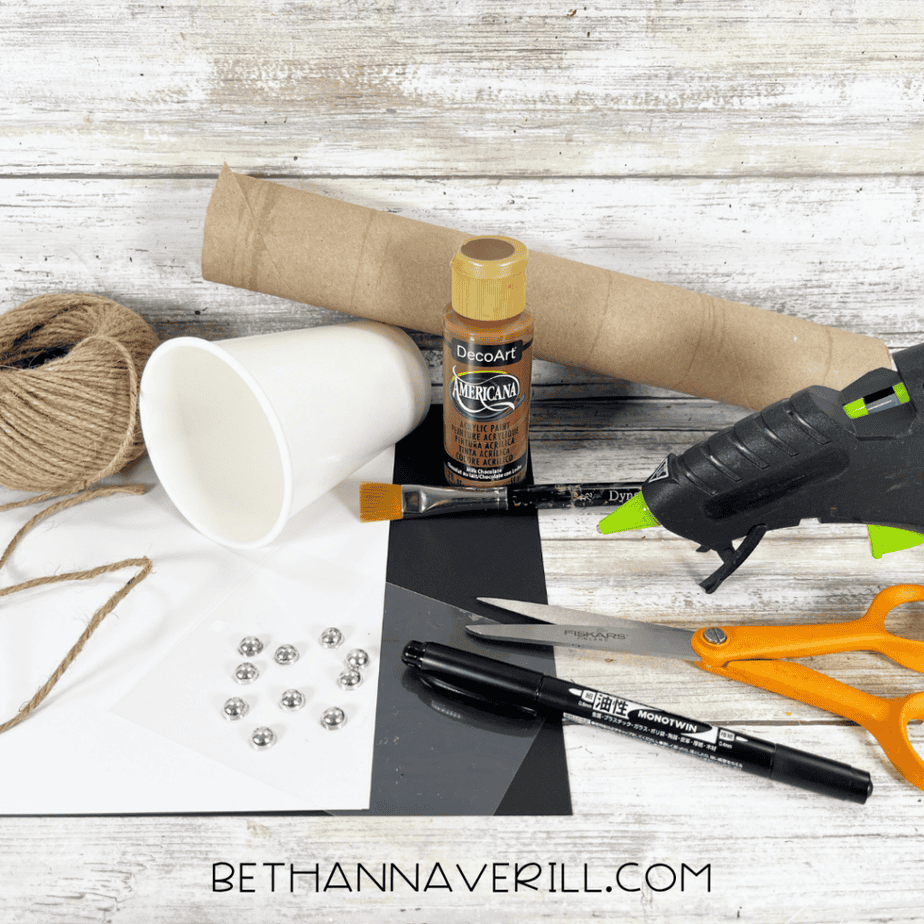 Craft supplies arranged on a rustic wood background: twine, white paper cup, cardboard tube, brown paint, paintbrush, hot glue gun, scissors, black paper, white paper, marker, and silver gems.