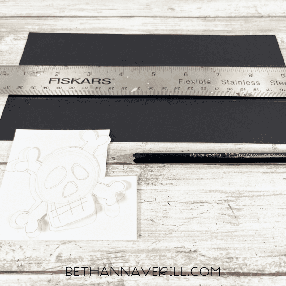 Black paper strips measured with a ruler, alongside a sketched skull-and-crossbones design on white paper.