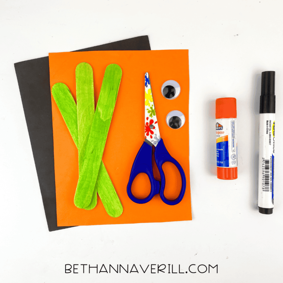 witch-craft-stick-project (2) Supplies for a witch popsicle stick craft including green painted craft sticks, orange and black cardstock, googly eyes, scissors, glue stick, and black marker on a white background.