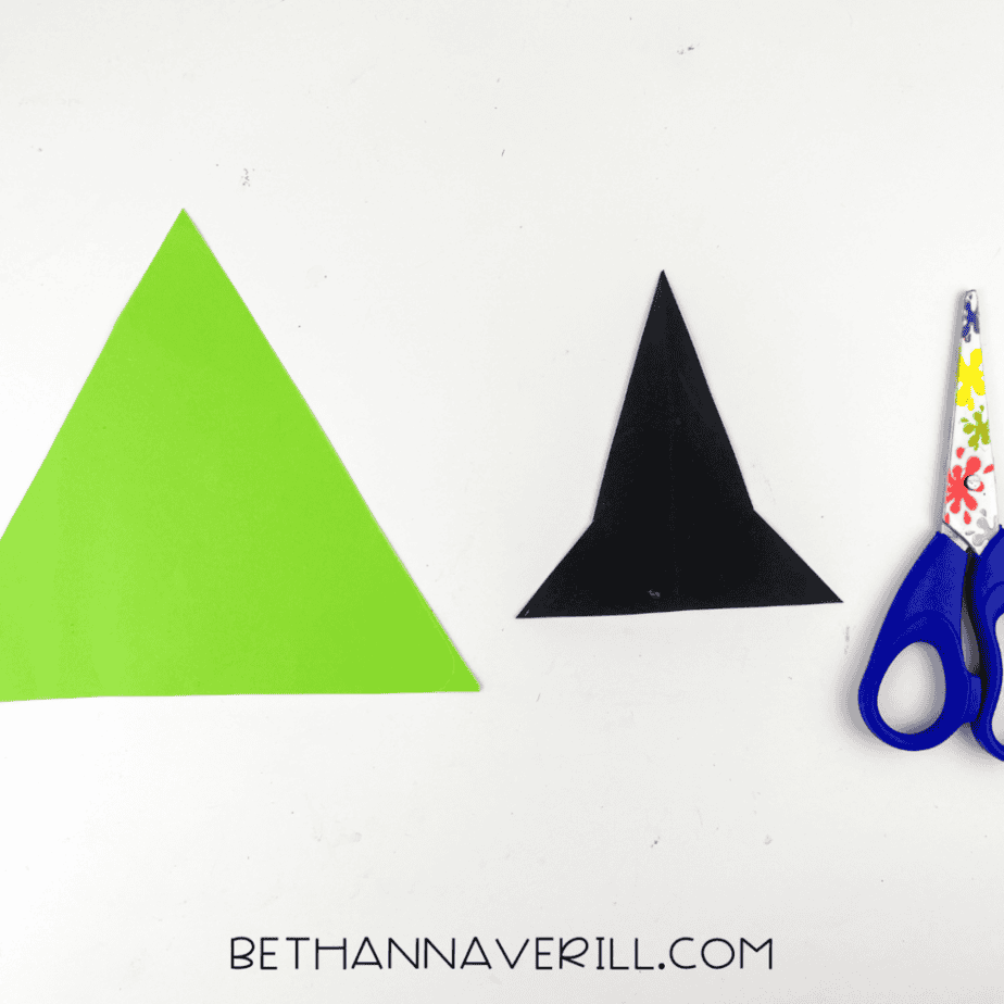 witch-craft-stick-project (3) Green and black paper triangles cut out for a witch craft project with scissors beside the paper shapes on a white table.