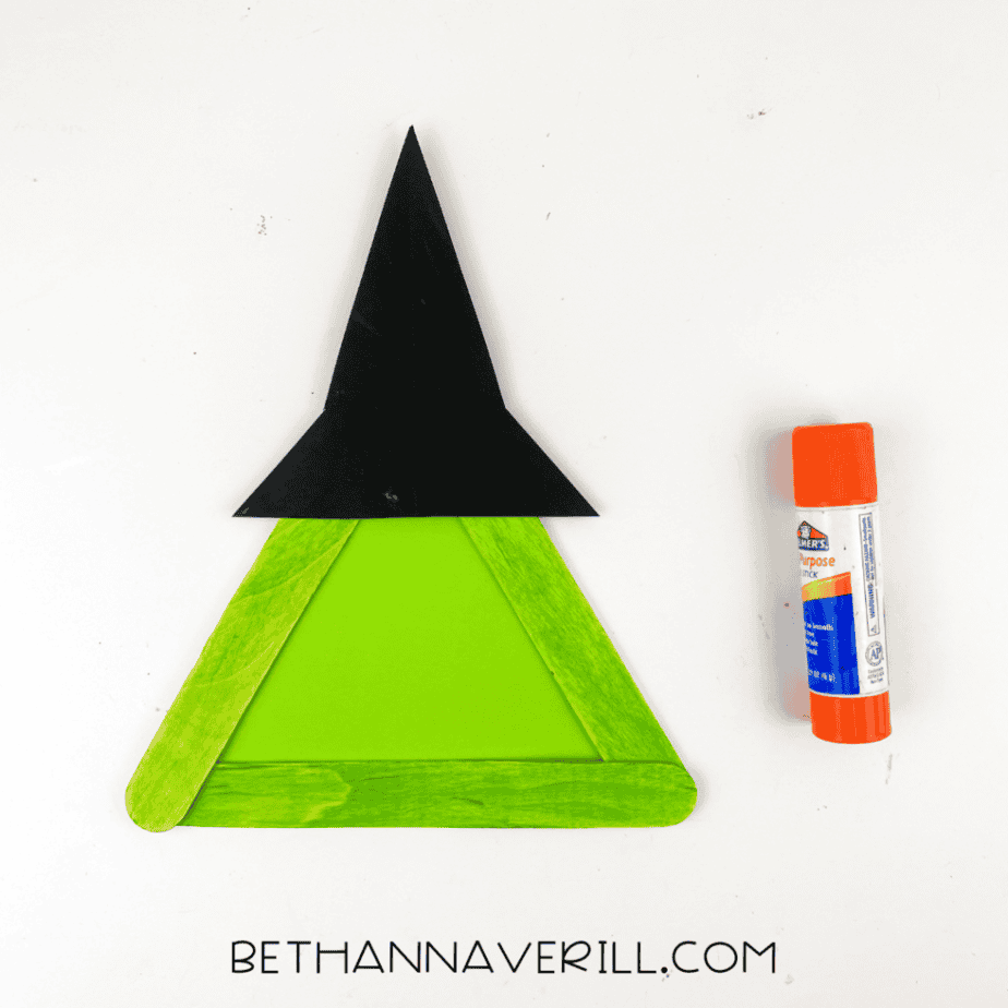witch-craft-stick-project (4) Green popsicle sticks glued into a triangle shape with green paper inside and a black paper witch hat attached to the top beside a glue stick.
