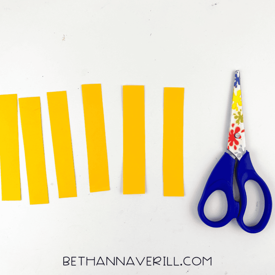 Yellow paper strips cut for witch hair placed beside blue handled scissors on a white background.