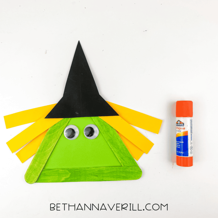 Witch popsicle stick craft in progress with yellow paper hair, googly eyes, black witch hat, and green triangle face beside a glue stick.