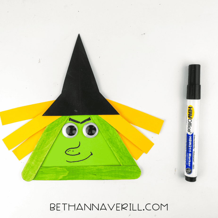 Finished witch popsicle stick craft with drawn face details, googly eyes, yellow paper hair, and black hat beside a black marker.