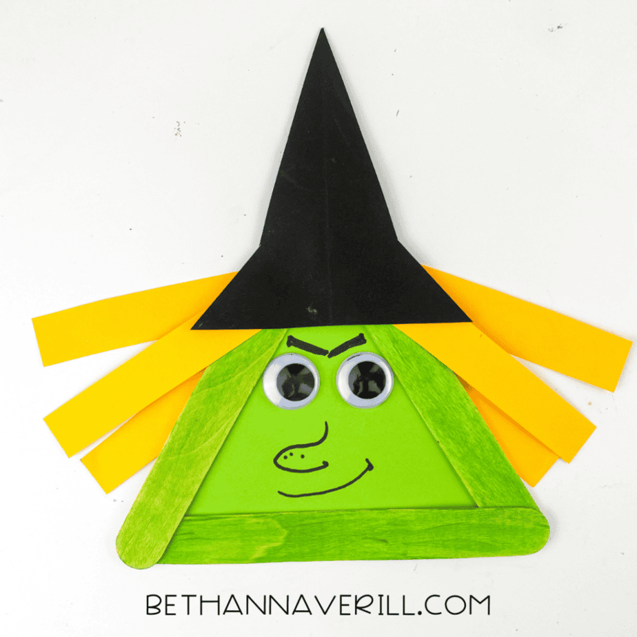 Close up of completed witch popsicle stick craft made with green craft sticks, googly eyes, yellow paper hair, and a black paper hat on a white background.
