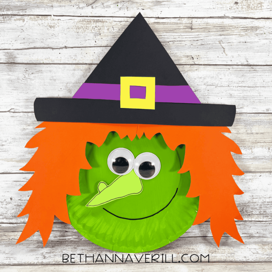 Finished green paper plate figure with orange jagged paper pieces, two googly eyes, a light green outlined paper shape, a curved black line, and a black pointed hat with purple and yellow paper details.