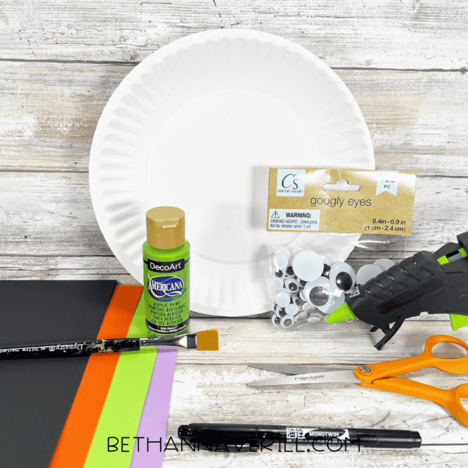 A white paper plate, green acrylic paint, a paintbrush, googly eyes, black, orange, green, and purple paper, a hot glue gun, orange-handled scissors, and a black marker arranged on a light wood surface.