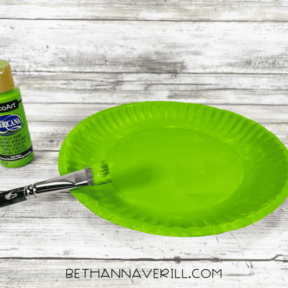 A paper plate painted bright green with a paintbrush beside it and a bottle of green acrylic paint partially visible.