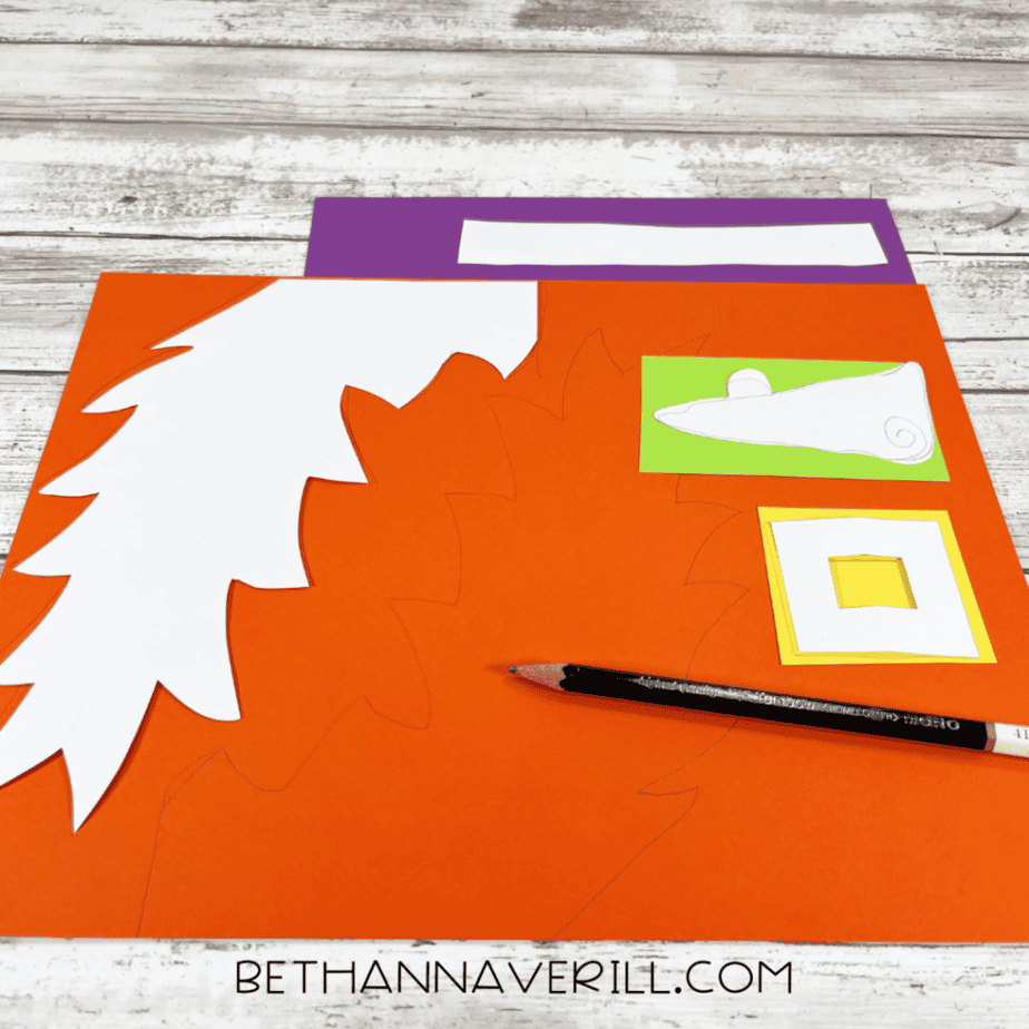 Orange, purple, green, yellow, and white paper pieces with drawn and cut shapes, plus a pencil, arranged on a light wood surface.