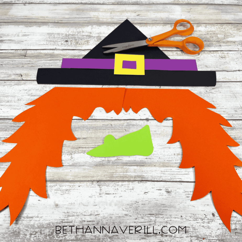 Cut paper pieces including a black triangular shape, black strip, purple strip, yellow square frame, two large orange jagged shapes, and a green curved shape, with orange-handled scissors above them.