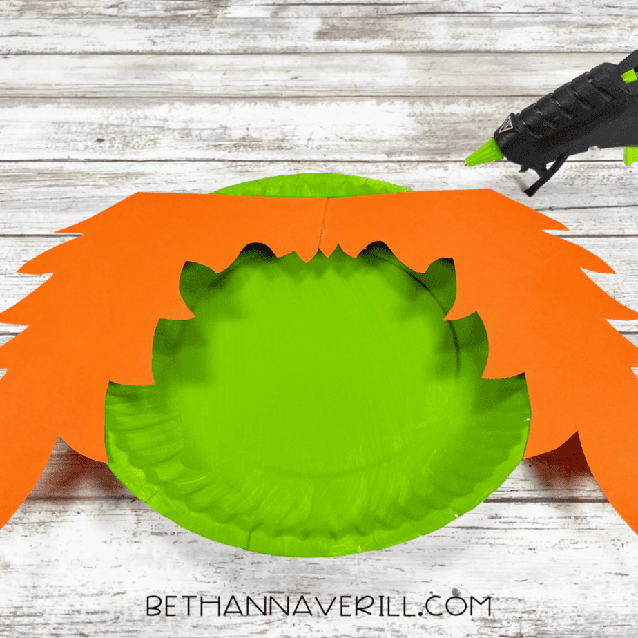 Two large orange jagged paper pieces positioned across the top of a green painted paper plate, with a hot glue gun in the upper right.