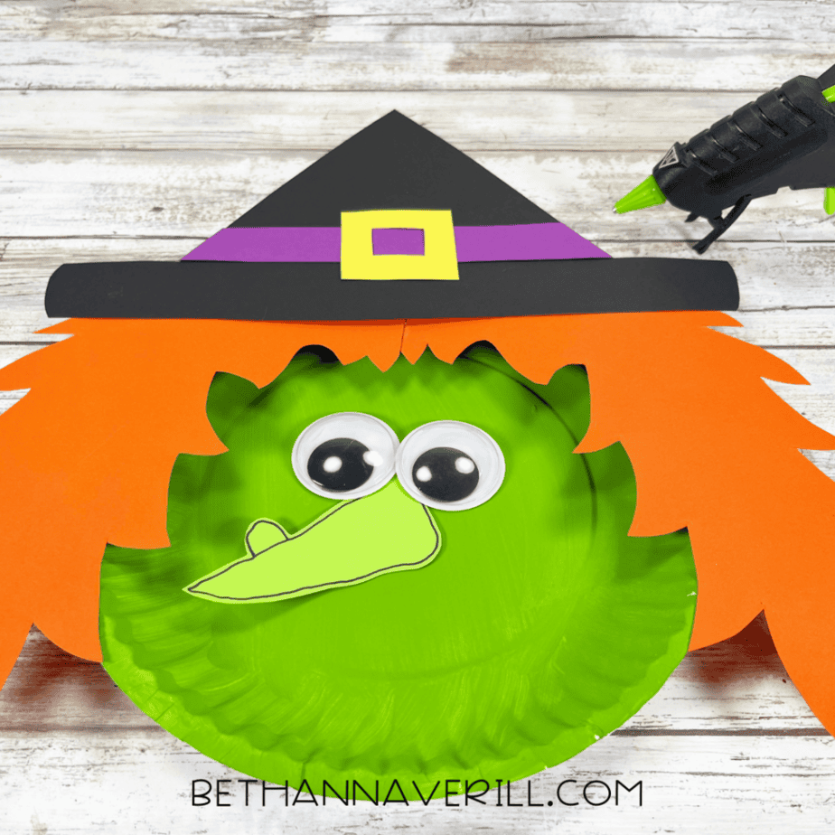 A green painted paper plate with orange jagged paper pieces, two googly eyes, a light green outlined paper shape, and a black paper hat with a purple band and yellow square detail.