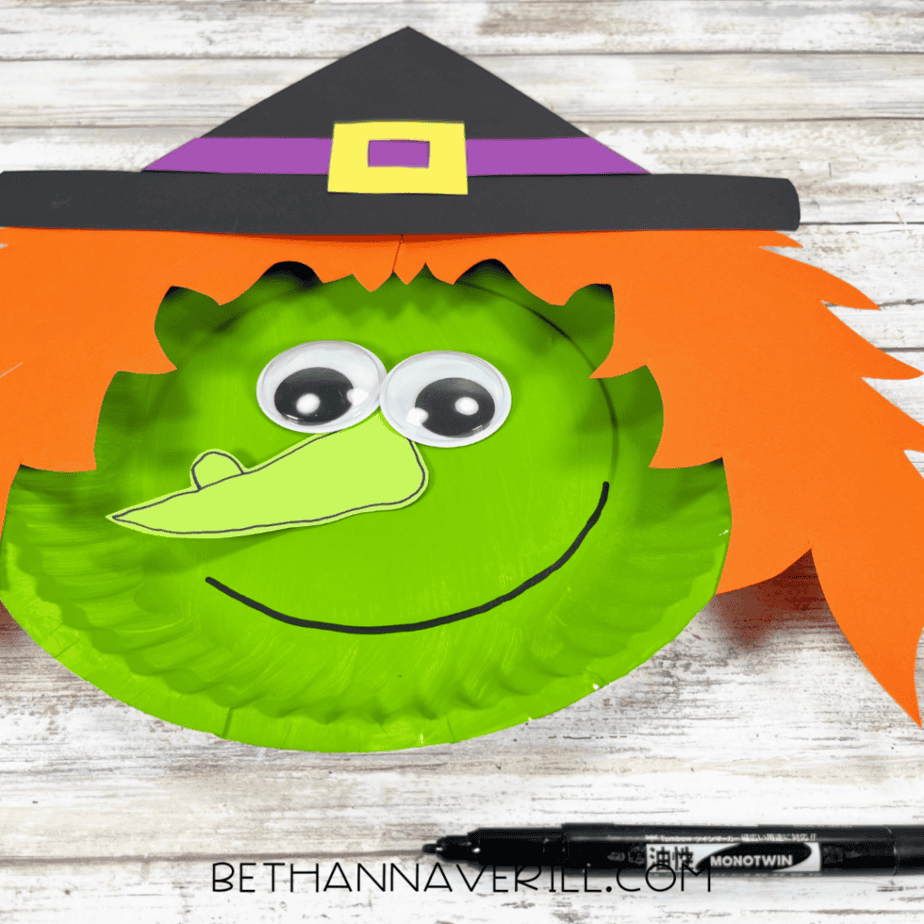 Close-up of the assembled paper plate figure with orange paper pieces, googly eyes, a light green outlined paper shape, a black hat with purple and yellow details, and a curved black line drawn across the lower part of the plate. A black marker is visible at the bottom.