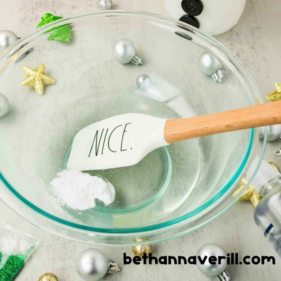 White powder and clear liquid sit in a glass mixing bowl beside a white spatula that reads “NICE.”. Silver ornaments and gold stars decorate the workspace. The watermark reads “bethannaverill.com”.