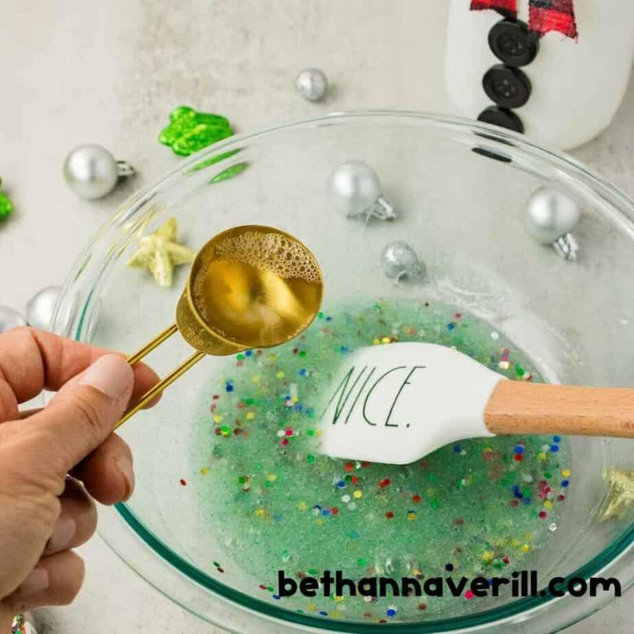A hand pours clear liquid from a gold measuring spoon into pale green Christmas Lights Slime filled with multicolored confetti. A white spatula reads “NICE.”. The watermark reads “bethannaverill.com”.