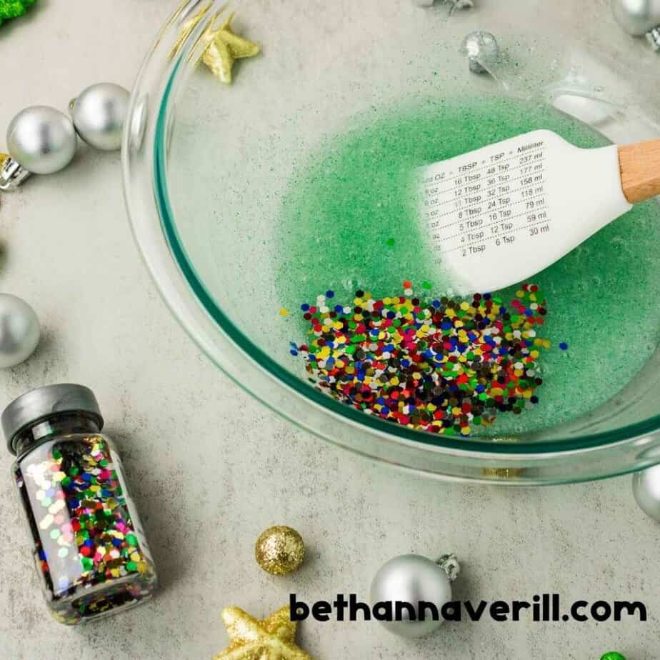 Multicolored hexagonal confetti rests on pale green slime inside a glass mixing bowl. A white spatula with a printed measurement conversion chart lies beside the mixture. The watermark reads “bethannaverill.com”.
