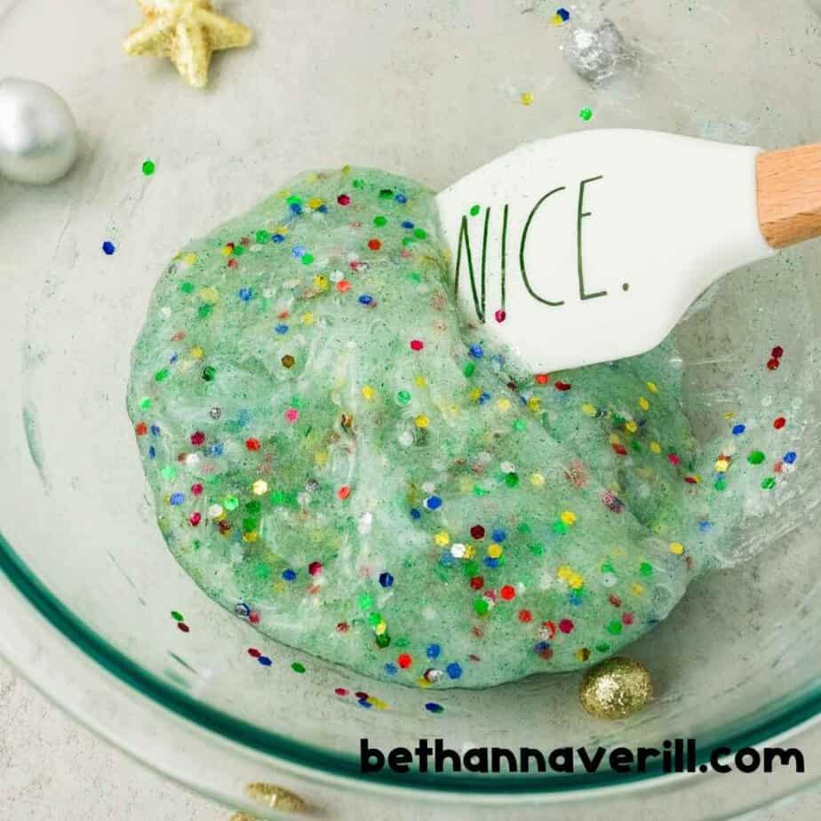 You may need to knead your Christmas Light slime by hand Thick pale green Christmas Lights Slime with multicolored confetti is folded inside a glass mixing bowl. A white spatula reads “NICE.”. The watermark reads “bethannaverill.com”.