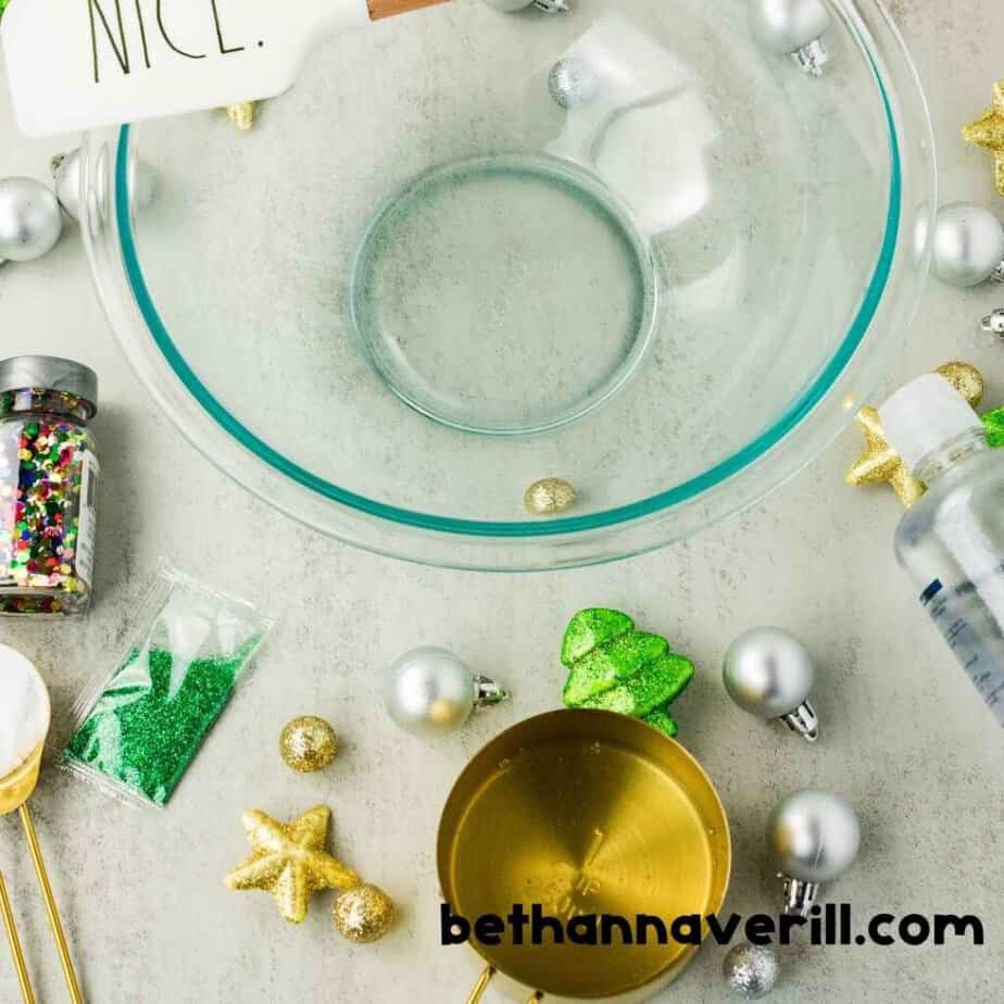 An empty clear glass mixing bowl sits among green glitter. Multicolored confetti. Silver ornaments. Gold stars. A measuring cup. And a bottle of clear liquid. The watermark reads “bethannaverill.com”.