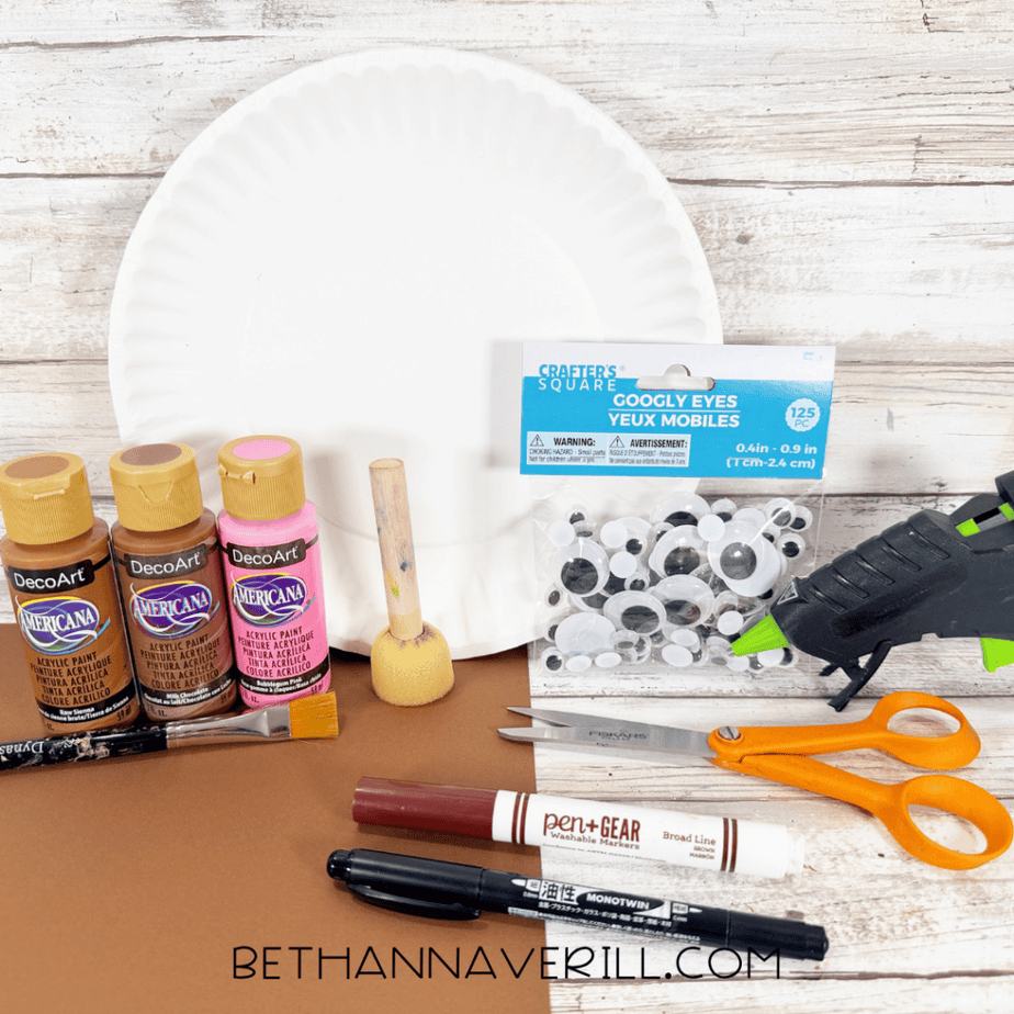 Supplies for an acorn paper plate craft, including a white paper plate, acrylic paints, googly eyes, paintbrushes, scissors, markers, brown paper, and a hot glue gun.