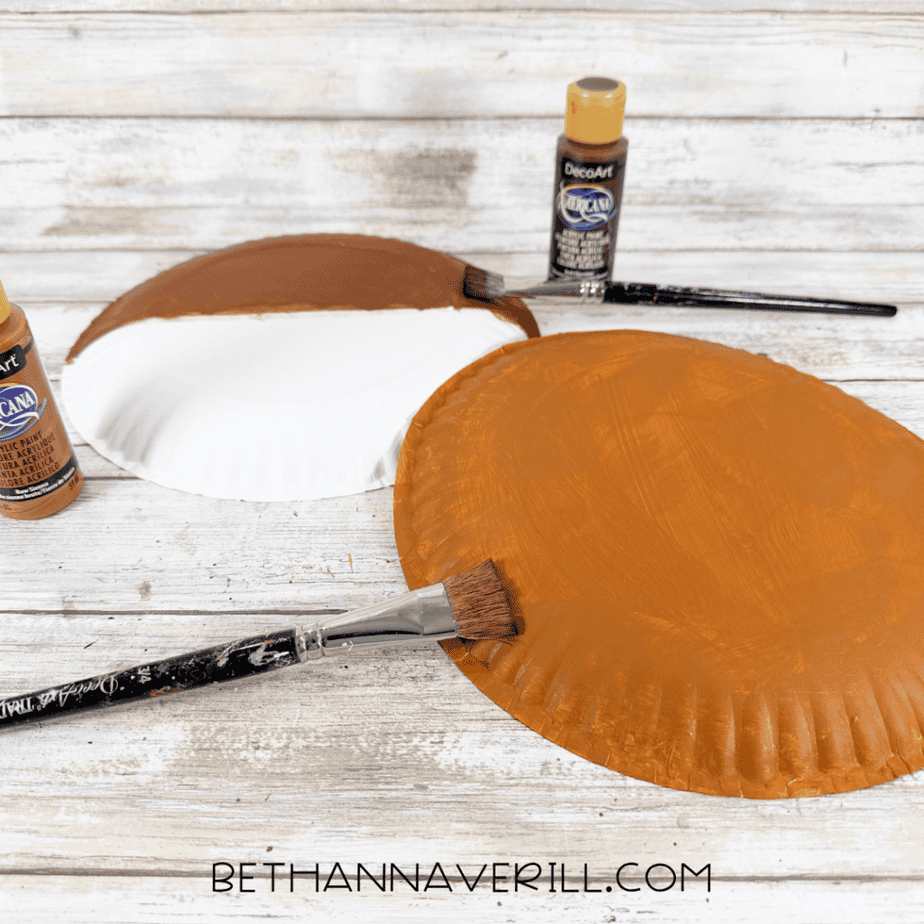 Paper plates being painted with brown and orange acrylic paint using paintbrushes on a white wooden surface.