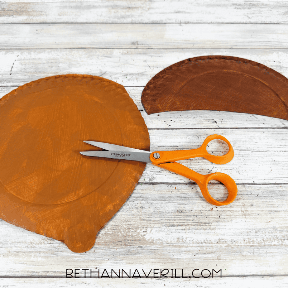 Orange painted paper plate cut into an acorn shape beside a brown painted semicircle and orange scissors.