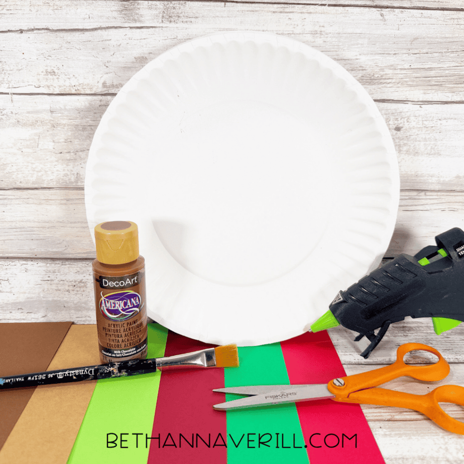 Supplies for an apple basket paper plate craft, including a white paper plate, brown acrylic paint, construction paper, paintbrush, scissors, and hot glue gun.