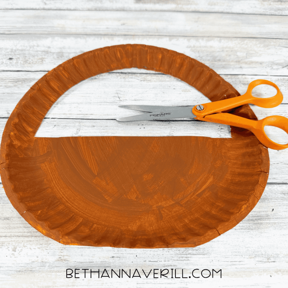 Brown paper plate with the upper center cut out to create a basket handle, with orange scissors beside it.