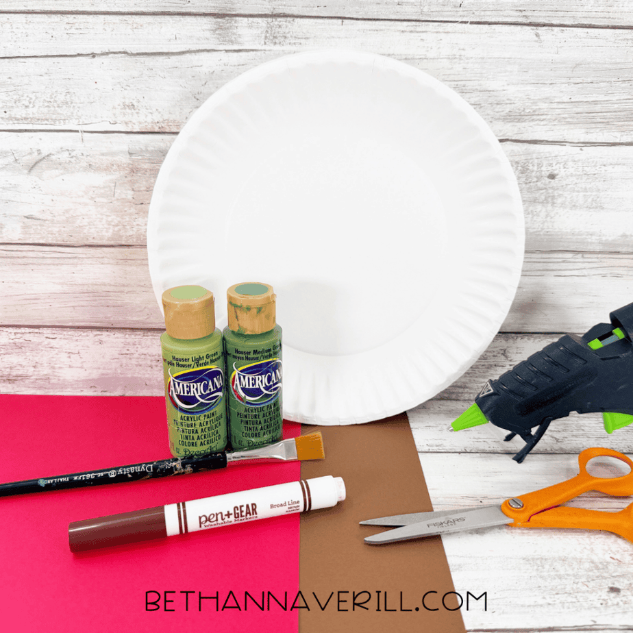 Supplies for an apple tree paper plate craft, including a white paper plate, green acrylic paints, red and brown construction paper, paintbrush, scissors, marker, and hot glue gun.