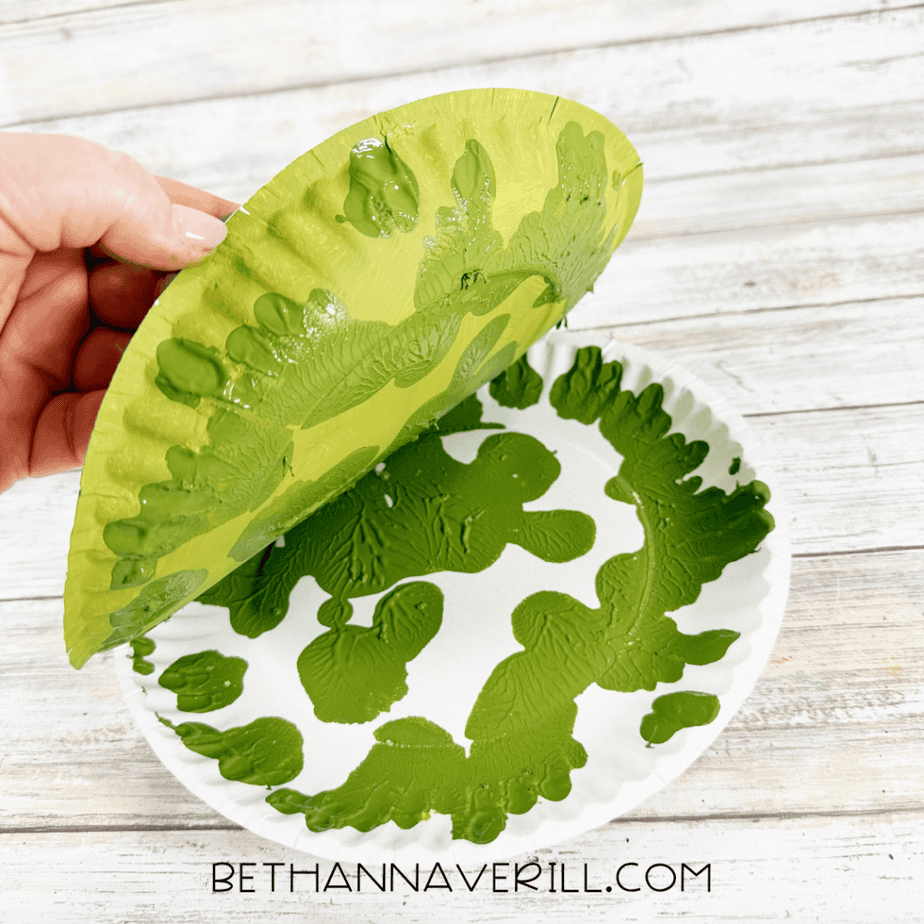 Hand lifting a light green painted paper plate from another plate to reveal dark green paint patterns.