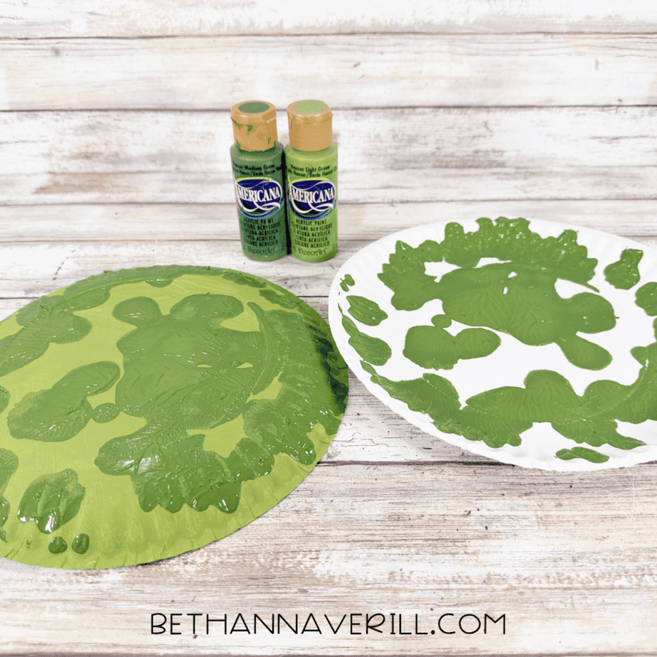 Two paper plates covered in light and dark green paint patterns beside bottles of green acrylic paint.