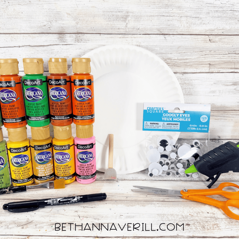 Supplies for an autumn leaves paper plate craft, including a white paper plate, colorful acrylic paints, googly eyes, paintbrush, sponge brush, scissors, marker, and hot glue gun.