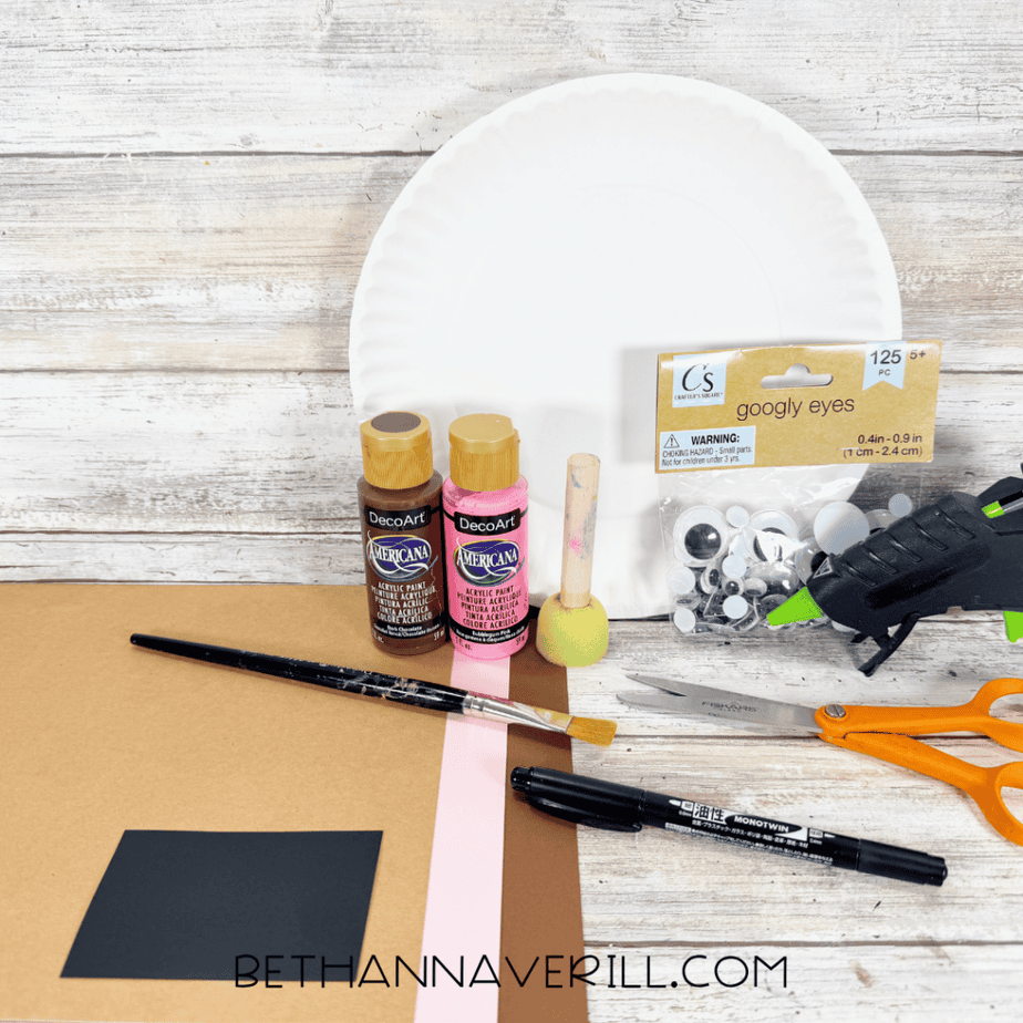 White paper plate with brown and pink paper, googly eyes, brown and pink paint, foam brush, paintbrush, scissors, black marker, and hot glue gun arranged on a white wood surface.