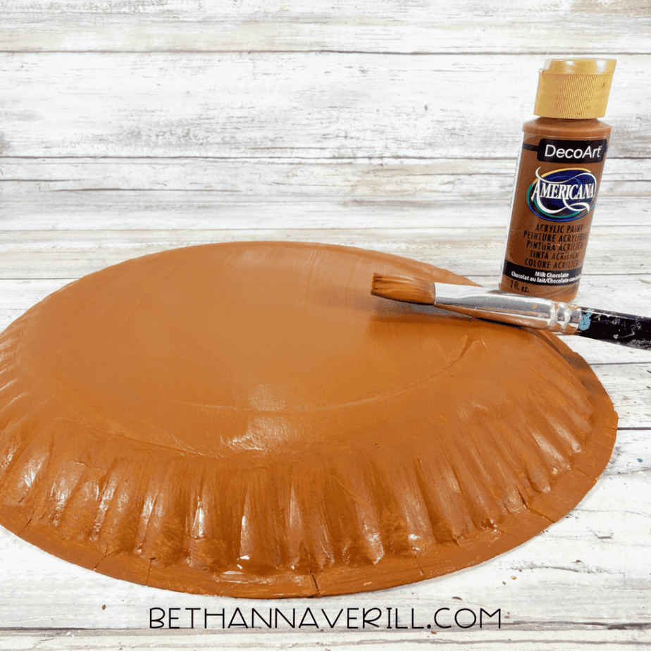 Paper plate painted brown with a paintbrush resting across it and a bottle of brown acrylic paint beside it.
