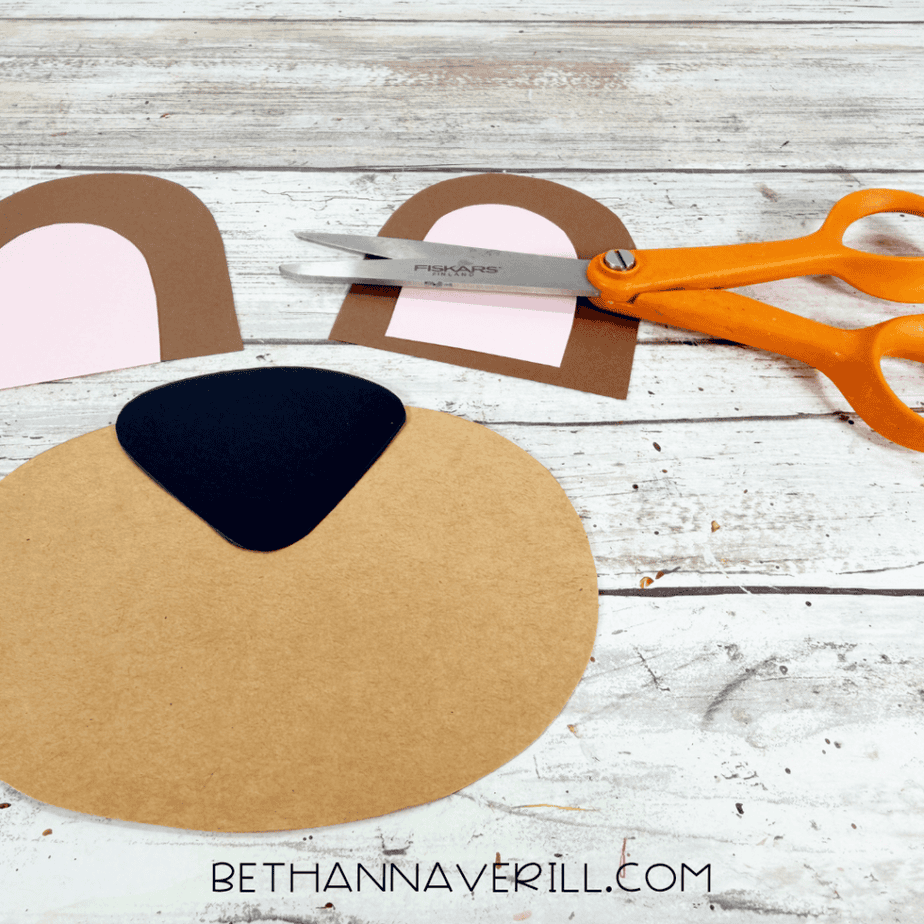 Cut paper pieces for a bear face, including a tan muzzle, black nose, and brown ears with pink centers, arranged beside orange scissors.