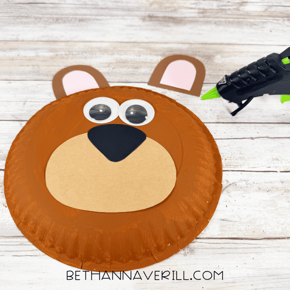 Brown painted paper plate with googly eyes, tan muzzle, black nose, and one ear attached while the second ear and a hot glue gun sit nearby.