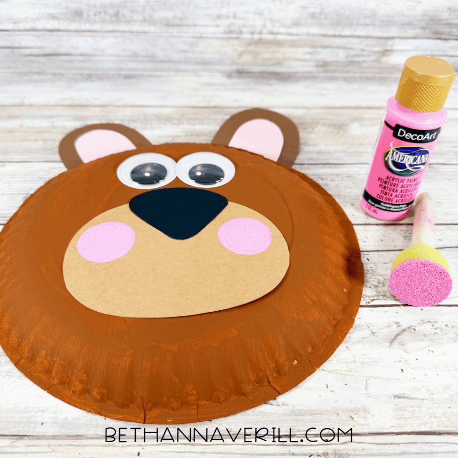 Brown paper plate bear with both ears attached, googly eyes, black nose, tan muzzle, and pink painted cheeks beside a bottle of pink paint and foam brush.