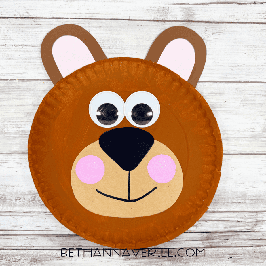 Finished brown paper plate bear craft with rounded ears, pink inner ears, large googly eyes, black nose, tan muzzle, pink cheeks, and a drawn smile.