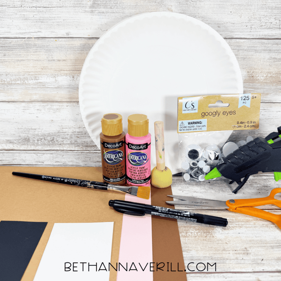 Paper plate craft supplies arranged on a white wood surface, including a white paper plate, brown and pink paint, googly eyes, paper, scissors, a paintbrush, marker, sponge brush, and hot glue gun.