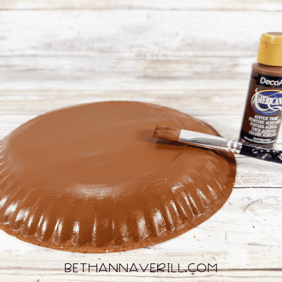 Brown acrylic paint being brushed onto the back of a paper plate on a white wood surface.