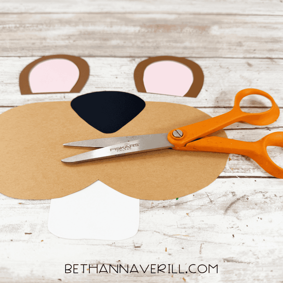 Cut paper pieces for the craft arranged on a white wood surface, including two ears, a tan muzzle, black nose, and white tooth piece.