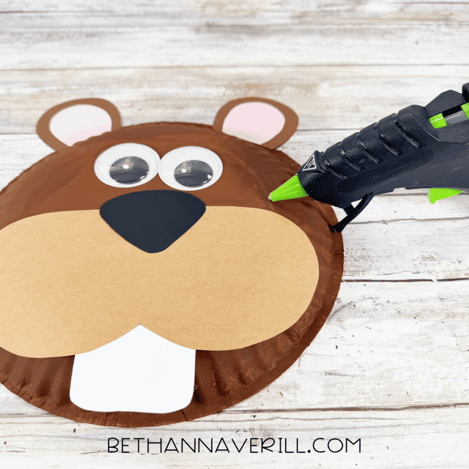 Partially assembled brown paper plate face with paper ears, googly eyes, tan muzzle, black nose, and white front teeth beside a hot glue gun.