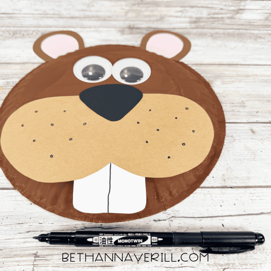 Brown paper plate beaver face with googly eyes, tan muzzle, black nose, front teeth, and small black dots drawn across the muzzle.