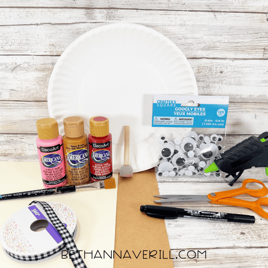 Supplies for a caramel apple paper plate craft, including a white paper plate, acrylic paints, googly eyes, craft ribbon, cardstock, paintbrush, scissors, marker, sponge brush, and hot glue gun.