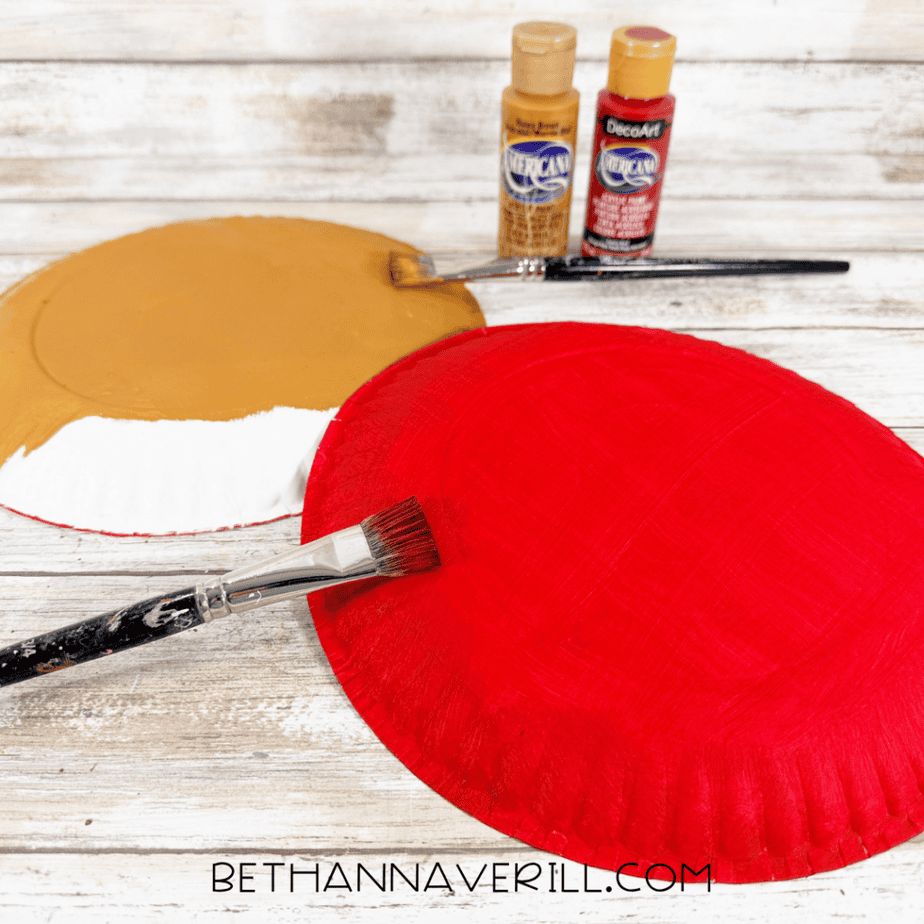 Paper plates being painted red and caramel brown with acrylic paint using flat paintbrushes.