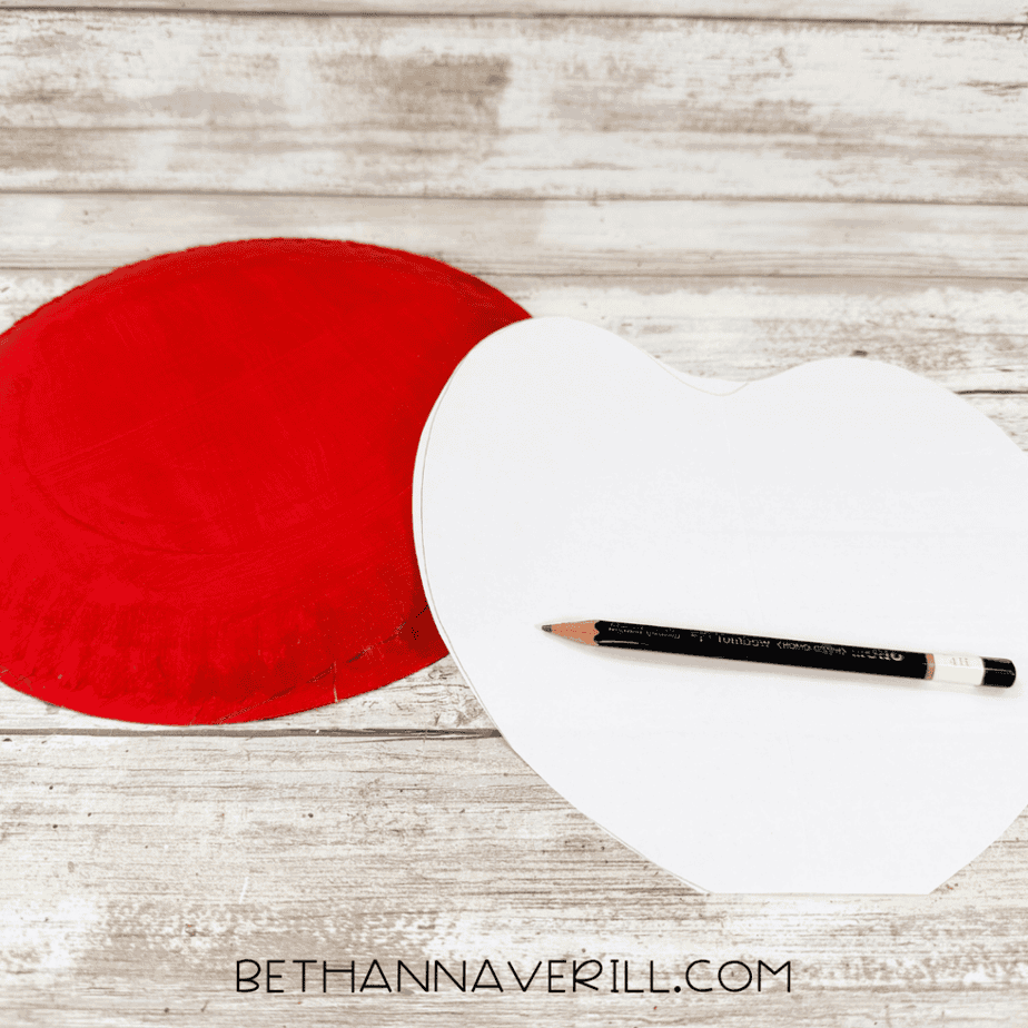 White paper apple-shaped template placed beside a red painted paper plate with a pencil for tracing.
